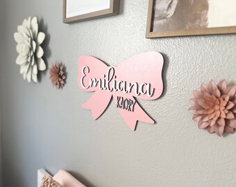 May include: A pink wooden bow-shaped wall decoration with the name "Emiliana" in white script. Below the name is "KAORY" in a smaller font. The bow is mounted on a gray wall, with paper flowers in shades of white and pink.