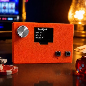 May include: A rectangular orange electronic device featuring a silver circular dial on the left, a small black screen displaying the text Blackjack, WIN: 24, BET: 5, DEALER: A, and two small black push buttons on the lower right side of the front panel.