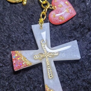 Könnte beinhalten: Ein goldfarbener Schlüsselanhänger mit einem Kreuzanhänger und einem Herzanhänger. Das Kreuz ist weiß mit goldenen Akzenten und Glitzer. Das Herz ist rosa mit Goldflocken. Der Schlüsselanhänger befindet sich auf einer schwarzen Oberfläche.