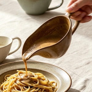 Puede incluir: Una salsera de cerámica marrón vierte una salsa espesa sobre un plato de espaguetis. Dos salseras de cerámica adicionales, una de color crema con motas y otra azul claro, se encuentran cerca sobre una superficie texturizada con un plato al fondo.