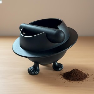Könnte beinhalten: Ein schwarzer Kaffeesatzbehälter mit einzigartigem Design. Der Behälter hat eine kreisförmige Basis mit drei fußartigen Füßen. Ein kleiner Haufen Kaffeesatz liegt neben dem Behälter auf einer Holzoberfläche. Der Behälter ist für einen Siebträger konzipiert.