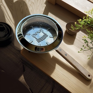 May include: A stainless steel cooking pan with a perforated interior featuring a castle design. The pan has a wooden handle and sits on a wooden cutting board. A small wooden bowl and greenery are also present.