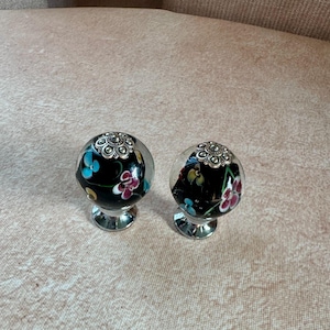 May include: Two decorative cabinet knobs. Each knob features a clear glass sphere with a black interior adorned with colorful floral designs. The knobs have a silver-colored metal base and a decorative silver top.