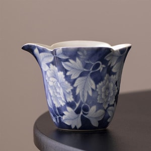 Può includere: Una tazza da tè in ceramica blu e bianca con un motivo floreale. La tazza ha un bordo smerlato e una base larga.