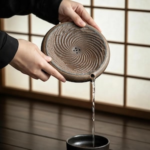 Könnte beinhalten: Ein rundes, braunes Keramik-Teigefäß mit texturiertem Spiralmuster auf der Oberfläche und einem kleinen Ausguss. Wasser fließt aus dem Ausguss in eine separate, schlichte schwarze Keramikschale. Das Gefäß hat eine kleine Gruppe von Abflusslöchern.