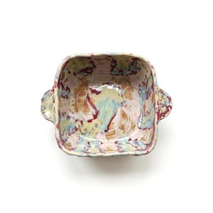 Puede incluir: Cuenco cuadrado de cerámica con dos asas. El cuenco presenta un diseño abstracto colorido con tonos de rosa, azul, amarillo y rojo. El interior y el exterior tienen un aspecto texturizado, lo que sugiere una calidad artesanal. El cuenco está sobre un fondo blanco.