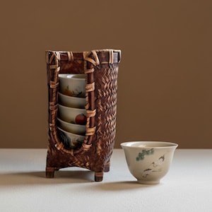 May include: A woven bamboo tea cup holder with a rectangular shape, holding several decorated tea cups. A single tea cup with a crane design sits to the right. The holder and cups are on a white surface against a brown background.