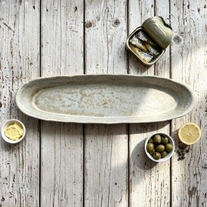 May include: An elongated, oval-shaped ceramic serving platter with a speckled, off-white and light brown glaze. The platter features a slightly uneven, organic rim and a textured surface, presented alongside a tin of sardines, olives, and lemon.