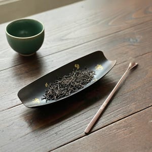 May include: A dark green ceramic tea bowl and a black serving tray with gold accents, filled with loose tea leaves. A silver tea spoon rests on a wooden surface. The tea set is designed for tea preparation and serving.