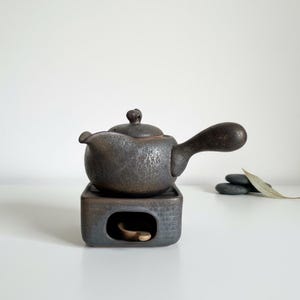 以下が含まれることがあります： 丸みを帯びたボディ、湾曲したハンドル、小さな蓋が付いた、濃い茶色のセラミックティーポット。 ティーポットは、小さな開口部のあるお揃いの正方形のベースに置かれています。 このセットは、お茶のサービス用に設計されています。