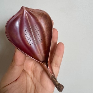 Puede incluir: Un colador de té de metal color cobre con forma de hoja o pétalo, con una punta afilada y una base estrecha. La superficie presenta un acabado texturizado y desgastado con un grupo central de pequeños orificios de drenaje circulares.