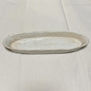 May include: An elongated oval ceramic tray with a textured, speckled beige and off-white glaze. The dish features a slightly raised, wavy rim and a smooth, shallow basin, showcasing a natural, earthy finish with subtle variations in color tone.