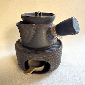 May include: A dark gray ceramic teapot set with a matching warmer and a small spoon. The teapot has a spout and a handle wrapped with twine. The warmer has a brick-like design with an opening for the spoon.