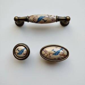 May include: A set of three cabinet handles: a long handle and two knobs. Each piece has a cream ceramic surface with a blue bird on a flowering branch design. The hardware has a bronze metal base. Ideal for drawers or cabinets.
