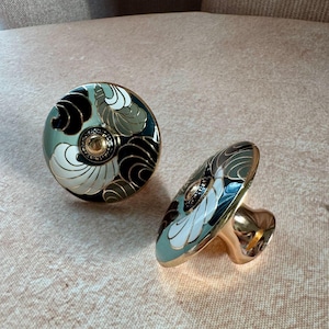 Pode incluir: Dois puxadores decorativos para armários. Cada puxador apresenta um design floral em preto, branco e verde oliva sobre um fundo verde pálido. Os puxadores têm uma base e um detalhe central dourados, adicionando um toque de elegância a qualquer móvel.