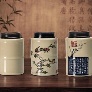 May include: Three ceramic tea canisters with black lids. The canisters are beige with bamboo-like detailing. One canister features a bird and floral design, another has a tree branch design, and the third has a calligraphy design.