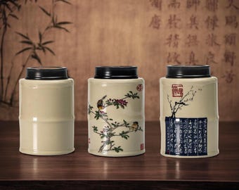 May include: Three ceramic tea canisters with black lids. The canisters are beige with bamboo-like detailing. One canister features a bird and floral design, another has a tree branch design, and the third has a calligraphy design.