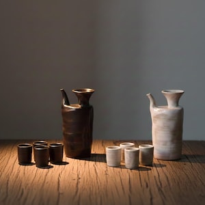 May include: Two ceramic sake sets, each consisting of a tall, slender carafe with a side spout and three matching small cups. One set is finished in a dark, matte brown glaze, while the other features a light, cream-coloured, textured glaze.