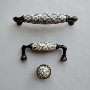 May include: A set of three cabinet hardware pieces: a long handle, a shorter handle, and a knob. The handles and knob feature a white floral pattern on a tan background, with dark bronze-coloured metal accents.