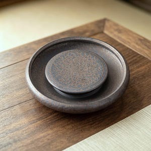 May include: A circular dark brown ceramic tea saucer with a raised central platform. The surface features a mottled, textured finish with a subtle, intricate embossed pattern covering the elevated center, creating a layered, dimensional appearance.