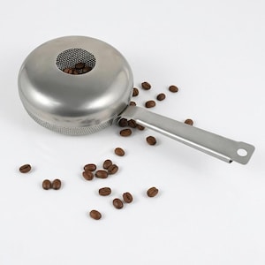 May include: A stainless steel coffee bean sifter with a round, perforated bowl and a flat handle featuring a circular hole at the end. The sifter is surrounded by scattered roasted brown coffee beans, with several beans visible inside the bowl.