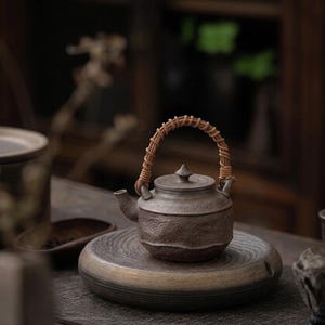 May include: A dark brown ceramic teapot with a textured, uneven surface and a small lid featuring a pointed knob. The teapot has a curved spout and a light brown woven rattan handle attached to two small loops on the top of the vessel body.