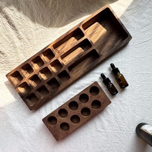 Op de afbeelding: Houten organizer set voor etherische olie. De set bevat een groot rechthoekig dienblad met meerdere compartimenten, een kleiner dienblad met ronde gaten en twee bruine glazen flesjes met zwarte druppelaars. Het hout heeft een diepe, donkerbruine kleur.