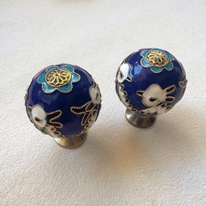 Könnte beinhalten: Zwei kugelförmige Schrankknöpfe mit tiefblauer Emaille-Oberfläche, verziert mit weißen Hasenmotiven und türkisfarbenen Blumenakzenten. Jeder Knopf ist mit feinem goldfarbenem Metalldraht umrandet und auf einem runden Metallsockel montiert.