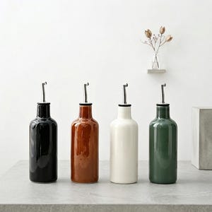Könnte beinhalten: Vier Keramik-Ölflaschen in Schwarz, Braun, Weiß und Grün, jede mit einem silbernen Ausgießer. Die Flaschen sind auf einer grauen Oberfläche angeordnet, mit minimalistischer Ästhetik. Im Hintergrund steht eine kleine Vase mit Trockenblumen.