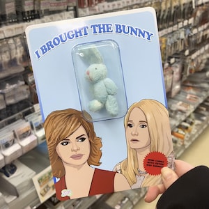 May include: A card with the text "I brought the bunny" and a cartoon image of two women. The card features a small blue bunny plush toy in a plastic case.