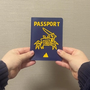Puede incluir: Funda de pasaporte azul marino con la palabra "PASSPORT" en letras amarillas. Un gráfico amarillo de una criatura estilizada está impreso debajo del texto. Un triángulo amarillo está en la parte inferior. La funda está sujeta por dos manos.