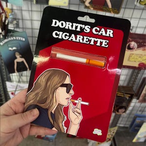 Puede incluir: Un artículo novedoso que presenta un cigarrillo en un estuche de plástico transparente, empaquetado en una tarjeta roja y negra. La tarjeta tiene el texto "DORIT'S CAR CIGARETTE" y una ilustración de una mujer fumando un cigarrillo.
