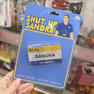 Può includere: Un cartellino identificativo blu e giallo con il testo "Hi! I'm SANDRA Team Member" su uno sfondo blu con il testo "SHUT UP SANDRA!" e un'immagine di un cartone animato di una donna che indossa una camicia blu con un cartellino identificativo. Il testo "For or What?" è in una nuvoletta sopra la testa della donna. Il testo "EMPLOYEE NAME BADGE" è in fondo alla carta.