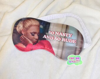 Può includere: Una maschera per dormire rosa e bianca con il viso di una donna e il testo "So Nasty and So Rude" stampato su di essa. La maschera ha un elastico bianco.