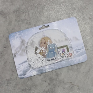 May include: A snow globe with a winter scene. Inside, a blonde woman and a blue bunny are surrounded by white beads. The base of the globe has the text "THE BUXX". The packaging features a winter landscape with snowflakes.