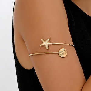 May include: Gold-toned arm cuff featuring a starfish and seashell design. The upper band has a starfish, and the lower band has a seashell. The jewelry is worn on the upper arm. The arm cuff is a beach-themed accessory.