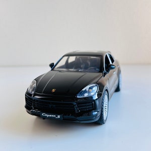 Puede incluir: Modelo negro de un SUV Porsche Cayenne S. El coche en miniatura presenta faros detallados, un acabado negro y el emblema "Cayenne S". El modelo se muestra sobre una superficie blanca.