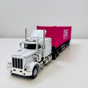 May include: A white and silver toy semi-truck with a pink container. The container has the word "ONE" printed on it. The truck has detailed features, including chrome accents and a black grill.
