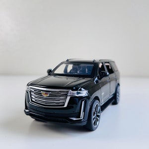 Puede incluir: Modelo de coche de juguete negro de un Cadillac Escalade SUV. El coche tiene una parrilla negra con un logotipo Cadillac cromado y ruedas negras.