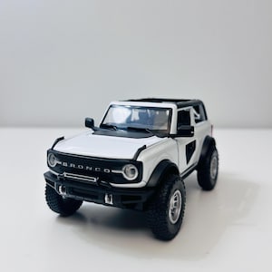 May include: A white and black Bronco model SUV with black tyres and the word "BRONCO" across the front. The model has a black roof and side mirrors. The vehicle is on a white surface.