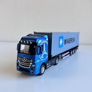 May include: A blue toy truck with a white container trailer. The trailer has the word "MAERSK" printed on the side.