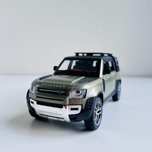 Puede incluir: Un coche de juguete Land Rover Defender verde con un portaequipajes de techo negro. El coche de juguete tiene un parachoques delantero plateado y la palabra "DEFENDER" está impresa en la parte delantera.