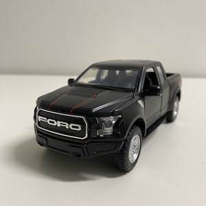Könnte beinhalten: Schwarzes Spielzeug-LKW-Modell eines Ford F-150 Raptor Pickups. Der LKW hat einen schwarzen und roten Streifen auf der Motorhaube und einen schwarzen Kühlergrill mit dem Wort "FORD" in silbernen Buchstaben.