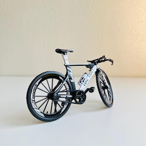 May include: A black and white time trial bicycle model with a water bottle holder. The bicycle has black wheels with white spokes and white lettering that says "Time Trial".