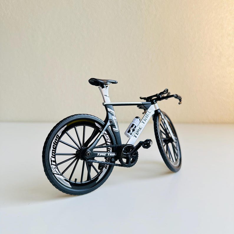 May include: A black and white time trial bicycle model with a water bottle holder. The bicycle has black wheels with white spokes and white lettering that says "Time Trial".