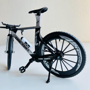 May include: A black and silver Time Trial bicycle with a water bottle holder. The bicycle has a black kickstand and black wheels with white lettering.