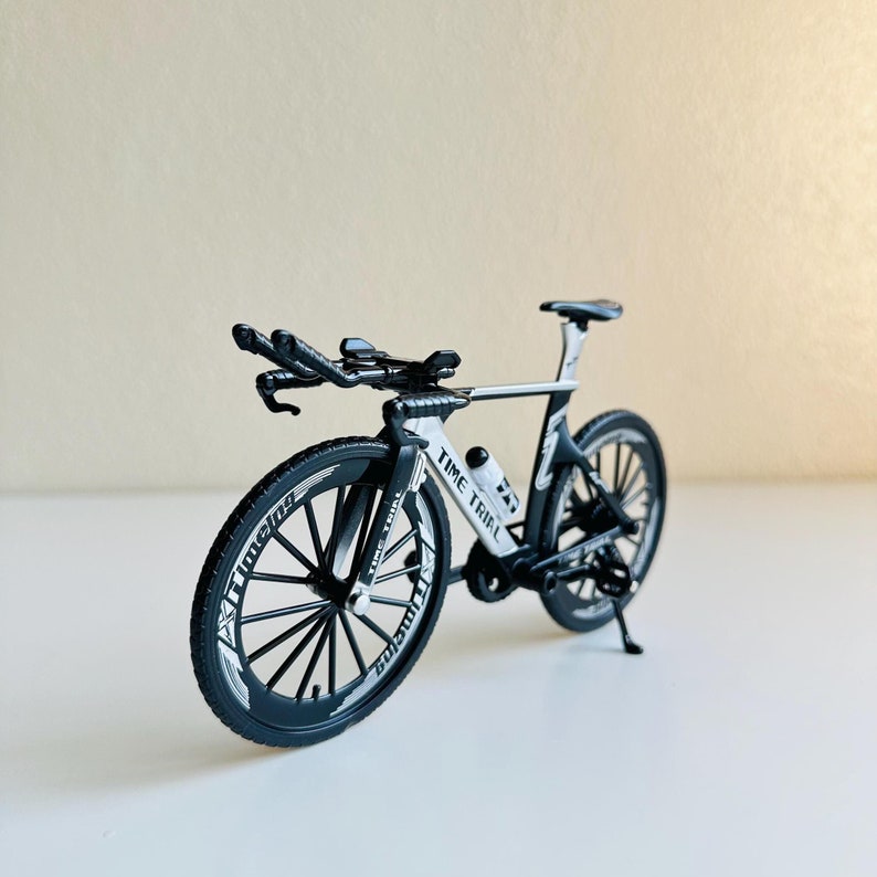 May include: A black and white time trial bicycle model with a black frame, white lettering, and black wheels. The bicycle has a water bottle holder and a kickstand.