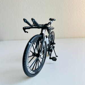 May include: A black toy bicycle with a silver frame and black wheels. The bicycle has a kickstand and a handlebar with two sets of handlebars.