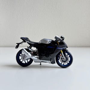 May include: A black and silver sport motorcycle model with blue accents. The miniature motorcycle has a detailed design, including a silver exhaust pipe, blue wheels, and a small windshield. The model is displayed on a white surface.