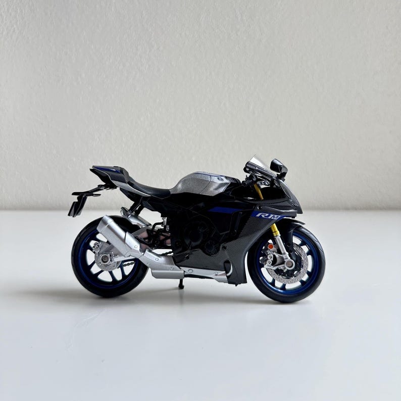 May include: A black and silver sport motorcycle model with blue accents. The miniature motorcycle has a detailed design, including a silver exhaust pipe, blue wheels, and a small windshield. The model is displayed on a white surface.